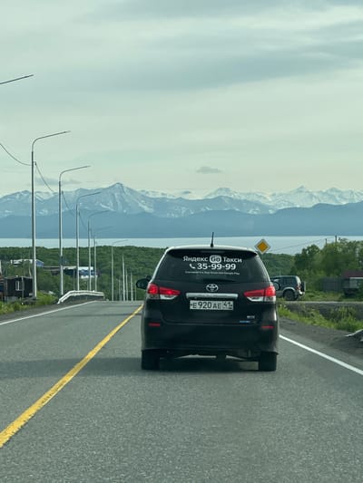 The image shows a rear view of a dark-colored SUV driving on a paved road in what appears to be a rural or suburban area. The SUV has a sticker on its rear windshield that says "Яндекс Go Такси" and a phone number. The license plate is visible and reads "E 920 AE 41 RUS". The road is marked with a yellow solid line on the left and a white solid line on the right. In the background, there are snow-capped mountains and a body of water, likely a bay or lake. The sky is overcast. There are several streetlights along the side of the road, and some houses and trees are visible. The location is identified as Krutoberegovy, Russia.