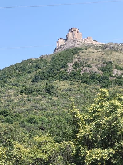 The image shows a stone church building situated atop a hill. The building appears to be ancient, with a brown, multi-tiered roof and stone walls. The hill is covered in dense green vegetation, with some rocky outcrops visible. The sky above is clear and blue. The foreground of the image shows more green foliage, with some lighter-colored trees. The scene is an outdoor, natural environment.