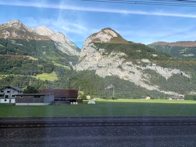 The image depicts a scenic view of the Swiss Alps, likely taken from a train moving parallel to a rural landscape. In the foreground, a section of train tracks with ballast is visible. Beyond the tracks, a bright green grassy field stretches towards a cluster of rural buildings. On the left, a modern gray residential building with balconies stands next to a long, dark wooden barn or storage structure. A row of white hay bales is neatly stacked in the field. In the middle ground, several farmhouses and other rural structures are scattered across the landscape, some with red roofs. The background is dominated by imposing, rocky mountains covered in dense green forests. The sky is a clear blue with a few wispy clouds. The image appears to be captured in the late afternoon or early evening, given the lighting and shadows. The location is identified as Schattdorf, Switzerland, which is consistent with the mountainous terrain and rural architecture.