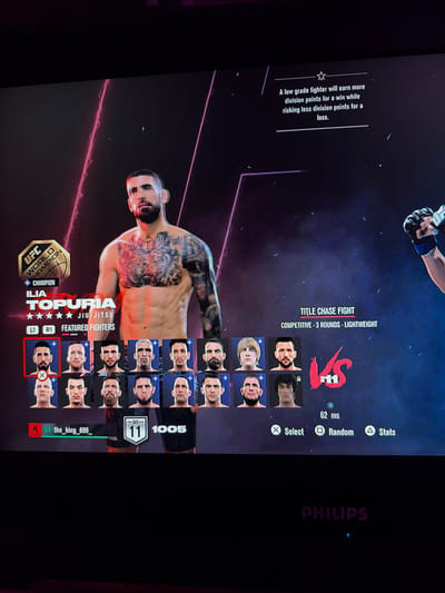 This is a photograph of a television screen displaying a UFC video game. The game interface shows a fighter named Ilia Topuria, who is the "UFC World Champion." Below his image, there is a selection of other fighter portraits, and the game is set up for a "TITLE CHASE FIGHT" which is "COMPETITIVE - 3 ROUNDS - LIGHTWEIGHT." The screen also shows player statistics, including a ping of "62 ms," and controller prompts for "Select," "Random," and "Stats." The bottom of the screen displays the "PHILIPS" logo, indicating the brand of television. There is no discernible information in the image that places the scene in Ludwigshafen am Rhein, Germany.