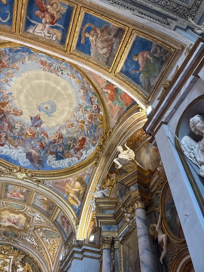 The image depicts the elaborate Baroque interior of the Chiesa del Gesù (Church of Jesus) in Foligno, Italy, viewed from below, looking upwards towards the ceiling and dome.
The central feature is a large, circular dome fresco portraying a celestial scene. A radiant light source emanates from the apex, surrounding a central divine figure, who is encircled by numerous ascending figures, including winged putti, angels, and saints on clouds, rendered in a vibrant palette of blues, reds, greens, and browns. This fresco is framed by an ornate gilded border.
Flanking the dome, the vaulted ceiling panels are also frescoed with various religious figures and scenes, set within intricate gilded stucco frames. Visible rectangular panels on the upper left depict individual figures, such as an angel on a cloud with a scroll and a white creature, and a draped figure also with a scroll. Other panels below these contain figures against a gold-toned background, enclosed by heavily gilded cartouches.
On the right side of the image, a substantial architectural pillar is visible. It features fluted, greenish-blue marble-effect columns with gilded Corinthian capitals, supporting an entablature adorned with rich gilded carvings. Multiple white stucco statues of winged figures are dynamically positioned on ledges along the pillar and archways. One prominent statue depicts a figure with wings, seemingly in motion, mounted on an architectural projection. Below it, another white stucco figure holds a lyre-like instrument. Further to the right and lower, a white stucco angel with wings is partially visible within a niche, holding a staff. The architectural elements are illuminated by modern spotlights affixed to the structure.
