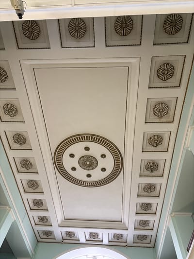 This image shows a close-up, low-angle view of an ornate, coffered ceiling. The ceiling is predominantly white with intricate decorative elements in a metallic, likely bronze or gold, finish. The coffering is divided into rectangular and square panels. Many of the square panels contain a raised, floral rosette design. The largest central panel features a large circular motif with concentric decorative rings and smaller floral accents. A light blue border runs along the top edge of the visible ceiling.
A small security camera is visible on the left side of the frame, attached to a white pipe or conduit. The camera is silver and black and appears to be mounted on a wall or structural element adjacent to the ceiling.
The scene appears to be indoors, likely in a building with classical or ornate architecture. The lighting is even and suggests it could be daytime, though no windows or natural light sources are directly visible, apart from a hint of a rounded window frame on the far right. The overall impression is one of grandeur and detailed craftsmanship. The location is identified as Luga, Russia. There is no text visible in the image.