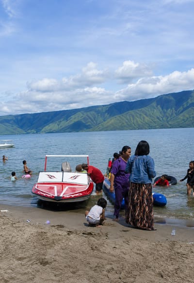 In the city of Situngkir, Indonesia, a group of people are enjoying a day at the beach. The scene depicts a body of water with several individuals, including children, wading and playing. A bright red and white motorboat is partially submerged near the shore. Several inflatable tubes are visible in the water, suggesting recreational activities. In the foreground, a child is crouched on the sand, appearing to play. In the midground, several adults are gathered near the boat and on the water's edge. The background features lush green mountains under a partly cloudy sky, indicating a natural, scenic landscape. The overall atmosphere suggests a casual, leisurely outing.