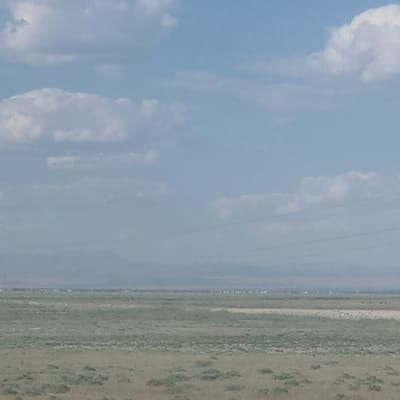 The image depicts a wide, flat landscape under a hazy sky with scattered clouds. The foreground consists of dry, sparse vegetation, appearing as patches of brown and green grass. In the middle ground, a band of lighter-colored earth runs horizontally across the scene, separating different areas of vegetation. Further in the distance, a faint, blurry line of structures suggests a settlement or town. A series of power lines are visible as thin dark lines stretching across the sky. The background is dominated by a hazy mountain range, almost lost in the atmospheric conditions. The overall impression is one of a dry, open expanse, possibly in a rural or semi-arid area. The location is identified as Shoytobe, Kazakhstan.