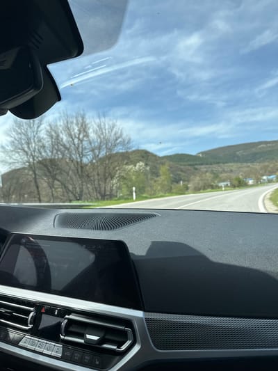 The image captures a view from inside a modern car, looking out through the windshield at a rural landscape. The perspective is from the passenger side, showcasing a significant portion of the car's dashboard, infotainment system, and the external scenery.
Inside the car, the dashboard features a textured, dark gray material, with a patterned trim panel running along the passenger side. A speaker grille is visible near the windshield. The infotainment system display shows "Music collection" and the time "11:46", along with "Previous track" indicating audio playback. Below the screen are climate control buttons and dials, including "AUTO", "MENU", "A/C", "MAX A/C", fan speed controls, temperature adjustments, and the hazard light button. The top of the windshield shows the housing for the rearview mirror or driver-assist cameras, with a dotted frit band along its edge.
Outside the vehicle, a clear blue sky with scattered white cirrus clouds dominates the upper portion of the view. A paved road, appearing to curve slightly to the right, stretches into the distance, flanked by green verges. Beyond the road, rolling hills or mountains are visible, covered in sparse, early spring vegetation and some bare-branched trees. The landscape suggests a journey through a scenic, perhaps mountainous, region. The bright sunlight and clear skies indicate a pleasant, sunny late morning.
The overall scene depicts a peaceful drive or stop in a natural setting. The visible text on the infotainment screen includes "Music collection", "11:46", "Previous track", and controls like "AUTO", "MENU", "A/C", "MAX A/C", and "OFF". The location context indicates this scene is set in Madrets, Bulgaria.