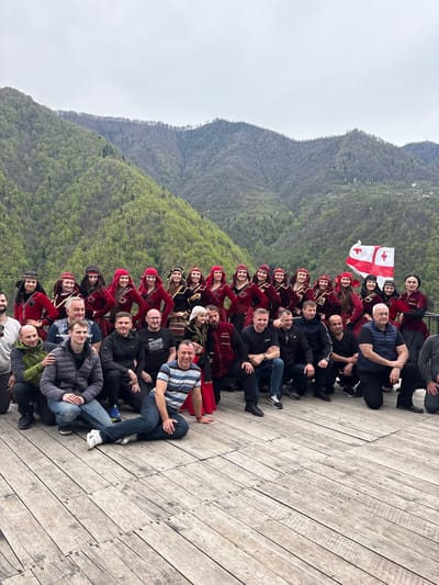 A group of approximately 40 people are seated on a wooden platform, posing for a photo. The majority of the group are wearing traditional Georgian clothing, specifically red velvet chokhas and hats. A Georgian flag is visible in the middle of the group. The platform overlooks a mountainside with a wooded area. The photograph was taken in Kveda Chkhutuneti, Georgia. The sky is overcast.
