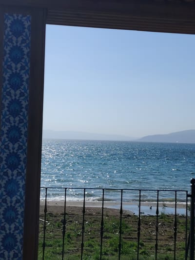 The image presents a picturesque view from a sheltered vantage point, likely a balcony or patio, overlooking a large body of water and distant hills.

On the left side, a vertical decorative panel features intricate blue and white patterns, resembling traditional Iznik tile work, which strongly suggests the location context of Iznik, Turkey. Above, a dark, horizontal wooden beam frames the top of the scene, implying a roof or overhead structure.

In the foreground, a black wrought-iron railing with ornate scroll designs stretches across the lower portion of the frame. Below the railing, a narrow strip of land shows patchy green grass and bare earth leading to a small, sandy or muddy beach where gentle waves lap at the shore.

The mid-ground is dominated by a vast expanse of shimmering blue water, likely Lake Iznik, with numerous bright reflections indicating sunlight glinting off its surface. In the distant background, multiple layers of hazy, light blue-grey hills or mountains recede into the horizon under a clear, pale blue sky.

The scene depicts a peaceful and serene environment during a bright, clear daytime, likely late morning or early afternoon, given the intensity of the sunlight. There are no people or specific activities taking place. No visible text is present in the image.