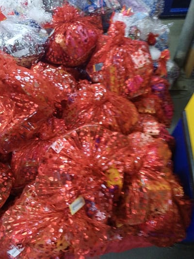 A stack of gift bags fills the frame, primarily red and silver. The red bags, adorned with a shimmering, reflective material, dominate the composition. Some bags have visible tags attached. The silver bags are clustered near the top left, appearing less voluminous than the red ones. The background is partially visible, showing a blue bin and a wall. The bags are tightly packed, creating a textured and festive impression.