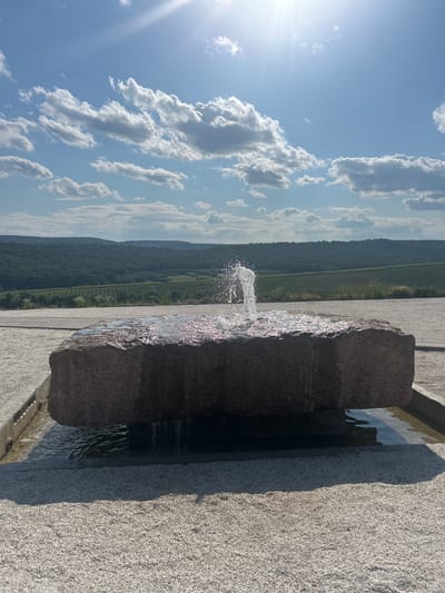 In the city of Ulmu, Moldova, on a bright sunny day with scattered clouds, a contemporary fountain features a large, rough-hewn stone as its centerpiece. Water spouts from the top of the stone, creating a dynamic splash that catches the sunlight. The stone rests in a shallow pool, with water gently overflowing its edges. The immediate surroundings are paved with small, light-colored gravel, creating a minimalist and clean aesthetic. Beyond the fountain area, a paved walkway leads towards a picturesque landscape. Rolling hills covered in dense green trees and vineyards stretch into the distance under a bright blue sky, with the sun creating a lens flare. The scene conveys a sense of tranquility and natural beauty integrated with modern design.
