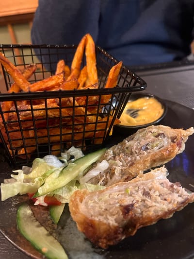 A plate presents two halves of a deep-fried spring roll, revealing a filling of shredded cabbage, meat, and vegetables. Sliced cucumber and lettuce garnish the plate. A small ramekin of orange dipping sauce sits beside the roll. Sweet potato fries, served in a small black metal basket, complete the dish.