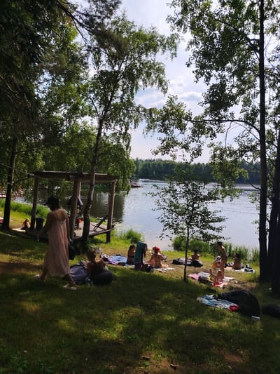 In the city of Vyltydor, Russia, a serene scene unfolds by a lake on a bright, sunny day. The image captures a group of people enjoying leisure time on the grassy shore, shaded by lush green trees.
In the foreground, a woman in a light pink dress walks away from the camera, her white sandals visible on the sun-dappled grass. To her left, another woman is seated on a mat, wearing a black bikini top and shorts. Further to the left, near the lake's edge, a woman with blonde hair sits on a wooden platform, wearing a black swimsuit.
The middle ground reveals more individuals scattered across the lawn, some lounging on blankets and inflatable cushions. A cluster of people are seen near the water, with one woman in a bikini standing and appearing to pour something from a bottle into a glass. Another woman, also in a bikini, is lying on her side on a blanket.
The background is dominated by the tranquil lake, its surface rippling slightly. A wooden structure, possibly a pier or a gazebo, is situated on the left side of the frame, adjacent to the grassy area. In the distance, across the lake, a dense forest line forms the horizon under a partly cloudy sky. There are some boats visible on the lake. The overall atmosphere is one of relaxation and outdoor recreation.