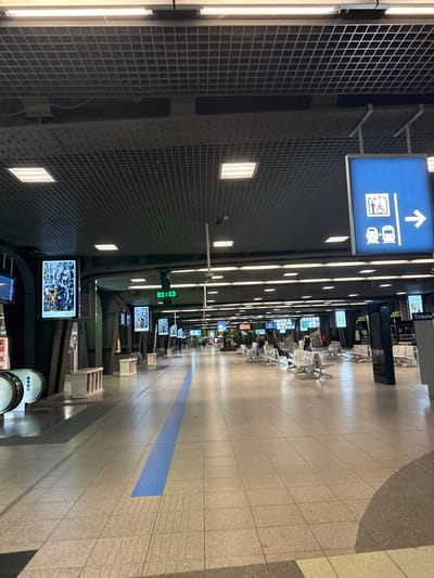 An indoor scene depicts a railway station hallway. A blue sign indicates train access. Along the right, seats face screens displaying information. A blue stripe runs the length of the floor. The ceiling features bright lights and a grid-like structure. A digital clock shows "01:13." The angle of the photograph suggests the image was taken by someone standing and turning the camera.