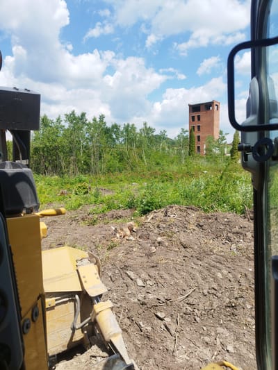 The image, taken from the perspective of a bulldozer cab in Rozdil, Ukraine, shows a partially cleared field with a brick tower in the background. The bulldozer's yellow blade and hydraulic arm are visible in the foreground, resting on disturbed earth. The field is overgrown with green vegetation and scattered tree stumps. The brick tower, appearing unfinished or abandoned, has multiple window openings and stands against a bright blue sky with scattered white clouds. A side mirror of the bulldozer reflects the sky and some of the surrounding greenery. The overall scene suggests ongoing construction or demolition work in a rural or semi-rural area.