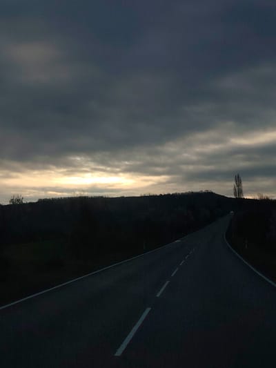 A ground-level, slightly low-angle shot shows a dark asphalt road with broken white lines curving uphill into the distance. The road is flanked on both sides by dark, shadowy vegetation. In the midground, the silhouetted horizon line is formed by a low hill covered in trees, punctuated by a single, tall, bare tree on the right side of the hill. Above the hill, the sky is filled with heavy, dark clouds, with a small band of lighter, golden light visible near the horizon. The overall impression is one of dusk or twilight. The location, Kesarevo, Bulgaria, implies a rural environment, which aligns with the landscape presented.