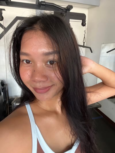 A close-up, selfie-style shot shows a woman with long, dark hair and brown eyes. She is wearing a light blue sports bra. Her skin appears slightly sweaty, and she is holding her left arm behind her head, with her hand near her hair. The background is blurred but suggests an indoor gym setting with black exercise equipment and a white wall. This scene is not specific to Cuyo, Philippines; it is a general image of a woman in a gym.