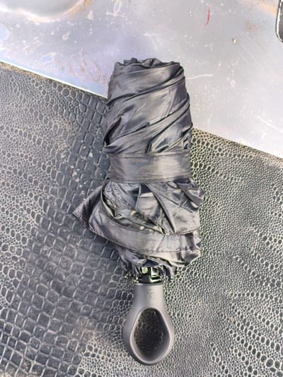 A black, folded umbrella lies on a dark, textured surface, possibly a car's interior or a similar material with a crocodile-like pattern. The umbrella is neatly wrapped and secured with a black band. Its handle is visible at the bottom of the frame, featuring a clover-like emblem. The background consists of a lighter, metallic-looking surface with scattered dirt or dust. The lighting suggests an indoor setting or an overcast day. There is no visible text in the image.