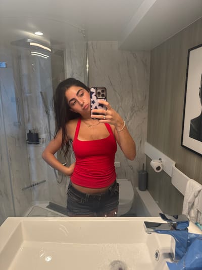 A female individual is depicted in a mirror reflection within a modern bathroom, located in Berry Islands, Bahamas. She has long dark hair and wears a red halter-neck tank top and dark denim cutoff shorts. Her left hand holds a smartphone, encased in a white and blue star-patterned cover, photographing her reflection. Multiple rings adorn her left hand, a silver bracelet is on her left wrist, and a gold chain necklace with a small pendant is visible. Her right hand rests on her hip, and her facial expression is a slight pout. The foreground shows a white rectangular sink with a chrome faucet, exhibiting water droplets. The bathroom's background features a glass-enclosed shower stall with white and gray marble-patterned walls, a rain showerhead, and a black soap dispenser. To the right, a partially visible white wall-mounted toilet, a dark gray toilet brush holder, a white wall-mounted shelf with a toilet paper roll, and a white towel on a rack are present. A framed black-and-white portrait of a profile hangs on the far right wall. Recessed overhead lighting is apparent, and the walls combine marble-patterned tile with light gray paneling.