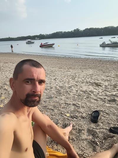 The image depicts a man sitting on a pebble beach with a body of water and a treeline in the background. He is shirtless and wearing black shorts. His right leg is bent at the knee, and his left leg is extended. A yellow towel is visible beneath him. In the sand to his right, there is a black flip-flop.
In the distance, on the water, there are several boats. One is a white speedboat with a red stripe, and another is a larger white boat. Further back, more boats are visible, some appearing to be barges. A person is standing in the shallow water near the shore. The sky is overcast with some patches of brighter sky, suggesting it might be late afternoon or early evening. The overall setting appears to be a beach or riverbank on a somewhat cloudy day. The man's expression is neutral. There is no visible text in the image.