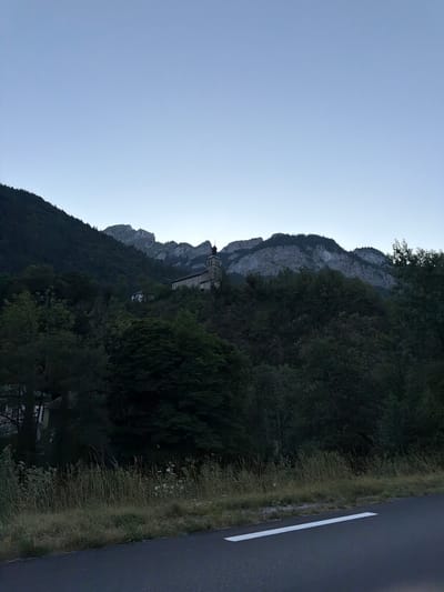 The image depicts a serene, twilight scene of a mountain landscape in La Baume, France. In the foreground, a grey asphalt road with a white line runs horizontally across the bottom of the frame, bordered by tall, dry grass and some wildflowers. The middle ground is dominated by a densely wooded hillside covered in various shades of green foliage, with a few scattered buildings visible amongst the trees. Perched higher on the hillside is a stone church with a tall steeple and a bell tower. Behind the church, imposing grey mountains with rugged peaks rise sharply against a pale blue, cloudless sky, indicative of early morning or late evening. The overall impression is one of natural beauty and tranquility.