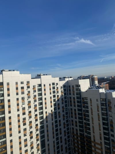 The image presents an urban landscape in Bittsa, Russia, captured from an elevated perspective. The upper portion of the frame is occupied by a clear, bright blue sky, featuring scattered, wispy white cirrus clouds, with a notable elongated cloud formation stretching across the upper right. The lower half displays a cluster of modern, high-rise residential buildings. These structures possess light-colored, possibly off-white, facades interspersed with dark-framed windows arranged in a grid pattern and vertical sections of light brown paneling. Numerous external air conditioning units are mounted on various building walls. Strong sunlight casts defined shadows from one building onto another, particularly on the right side of the image, obscuring parts of a facade in shade. Building rooftops are flat and feature visible mechanical equipment. In the far distance, additional city infrastructure is visible, alongside a segment of what appears to be a forested area. Some distant rooftops exhibit traces of snow. No human figures, discernible actions, or interactions are present in the scene.