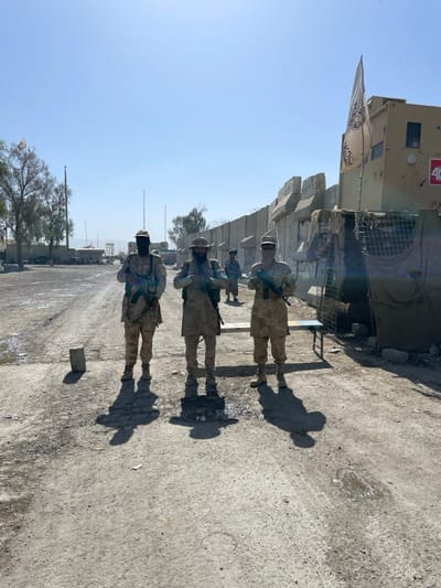 The image depicts a dusty, open-air scene in Spin Buldak, Afghanistan, characterized by a military or security presence under a clear, bright sky.
In the foreground, three men stand facing the viewer, positioned on an unpaved, uneven dirt and gravel surface. All three are dressed in camouflage uniforms, which appear to be light tan or desert-patterned. The man on the left wears a tan cap and a black balaclava covering his face, holding a rifle (AK-variant) vertically. The central figure wears a tan bucket hat and has a dark beard, also holding an AK-variant rifle. The man on the right wears a tan cap and a light-colored face covering, similarly armed with an AK-variant rifle. All three cast distinct, elongated shadows to their right, indicating strong overhead sunlight. Near the feet of the leftmost man is a rectangular concrete block. A small, blue and white striped metal bench is situated to the right of the foreground figures.
Behind the figures, a long wall composed of large, modular concrete barriers or Hesco barriers extends horizontally across the midground. Barbed wire is visible along the top of this wall in sections. To the right, immediately behind this wall, a fortified structure or vehicle, possibly a tan armored personnel carrier or bunker, is visible. A white flag with black Arabic script, identifiable as a Taliban flag, flies from this structure. A red "4G" decal is also present on its side. Further down the dirt path in the midground, two additional individuals in similar attire are partially visible, one standing near a parked vehicle.
The background shows sparse, dry trees on the left and more structures and distant terrain. The horizon reveals faint hills or mountains under the unbroken blue sky. The overall environment appears arid and operational, possibly a checkpoint or military outpost.