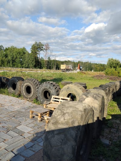 In Novoselytsia, Ukraine, on a sunny day with scattered clouds, a construction site is depicted. The foreground features a paved area with large, heavy-duty tires arranged in a row, suggesting they might be repurposed for an industrial or artistic installation. Two wooden pallets have been assembled into a simple table and seating area, placed on the paving stones. In the background, a dump truck and an orange excavator are positioned on a grassy slope, indicating ongoing construction or demolition work. A line of trees borders the area, with a glimpse of a wooden fence in the mid-ground. The scene is devoid of any people.