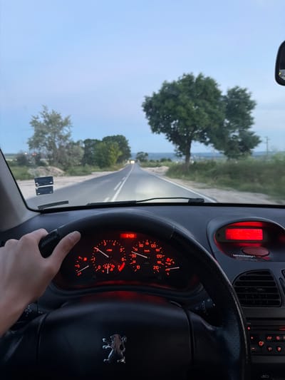The image is taken from inside a car, looking forward through the windshield. The driver's left hand is on the steering wheel. The dashboard displays the speedometer, which shows approximately 40 km/h, and other indicators such as fuel level and temperature. The car is on a paved road, with fields and trees on either side. In the distance, the road recedes towards the horizon, with the headlights of an oncoming vehicle visible. The sky is overcast, suggesting it is either dawn or dusk. The scene is likely located in or around the village of Ignatievo, Bulgaria, given the context.