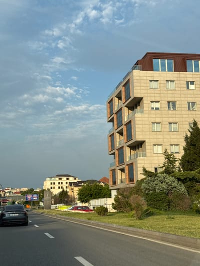 This is a media file depicting a street scene in Baku, Azerbaijan, likely during the late afternoon or early evening given the lighting. The image is oriented vertically, with the sky occupying the left and bottom portions and the cityscape and road filling the right and top.
The primary subject is a modern, multi-story building with a distinctive architectural style, featuring a dark brown facade on the upper section and a lighter, textured beige facade below. This lower section has prominent, recessed balconies with glass railings, creating a tiered effect. Adjacent to the building is a well-maintained green lawn and several trees, including evergreens and some with fuller foliage.
To the right of the building, a paved road with white lane markings is visible, suggesting it's a public thoroughfare. A black car is driving on this road, and its license plate is partially visible, appearing to read "90 TB 692". Further down the road, other buildings and some advertisements are visible, including a billboard with text that is difficult to read clearly.
The sky is partly cloudy, with wispy white clouds spread across a pale blue background. The overall mood of the image is urban and contemporary, showcasing a blend of modern architecture and greenery. There are no people clearly visible in the scene. The weather appears to be clear, but with some overcast conditions contributing to the muted light. The location cues, such as the architecture and the style of the road, align with an urban setting in Azerbaijan.