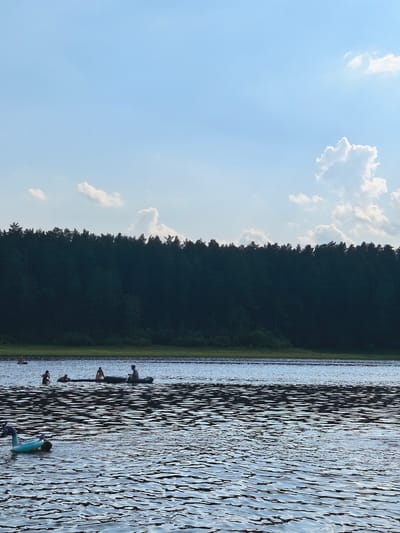 This media file captures a serene summer scene on a lake in Sysert, Russia. The image is dominated by the calm, rippling water, reflecting the clear blue sky dotted with fluffy white clouds. In the distance, a dense line of dark green trees forms a natural backdrop to the tranquil setting. The time of day appears to be late afternoon or early evening, as indicated by the soft lighting and the slight haze in the air.
Several individuals are enjoying recreational activities on the water. In the foreground, an inflatable blue toy, possibly a pool float, is partially submerged. Further out on the lake, a group of people are engaged in various water activities. One group is in a canoe, with at least two individuals paddling, while another person sits in the boat. Nearby, other individuals are swimming or wading in the water. The atmosphere seems relaxed and pleasant, suggesting a leisurely day spent outdoors. The overall impression is one of peaceful enjoyment of nature's beauty. There is no visible text in the image.