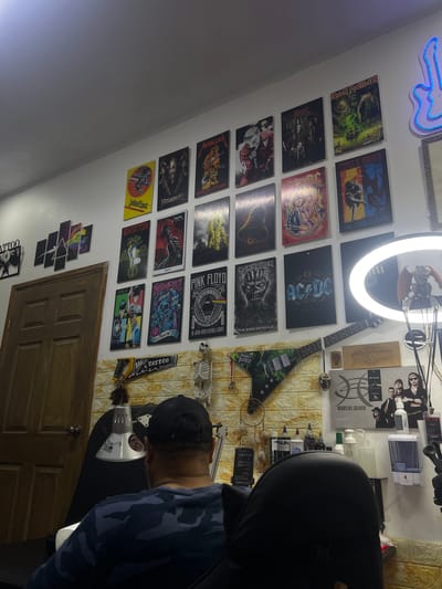 The image shows the interior of a room, possibly a tattoo studio, in Santa Rosa de Copán, Honduras, judging by the tattoo-related items and decor. A man wearing a dark-colored baseball cap and a dark blue camouflage-patterned shirt is leaning over a desk. On the wall, there is a collection of framed posters featuring various rock and metal bands, including Iron Maiden, Metallica, AC/DC, and Pink Floyd. Also visible are a neon sign, a ring light, a decorative guitar, and a mannequin skeleton. The background includes a yellow brick-patterned wall and a wooden door. A variety of tattooing supplies and equipment are arranged on the desk, including a lamp and various bottles, likely containing tattoo inks and solutions.