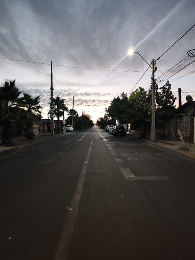 A street scene under a cloudy sky, possibly at dusk. The road stretches into the distance with a solid white line down the middle. Buildings and parked cars line the sides. Trees with leafy foliage are present on one side, while palm trees stand on the other. Power lines crisscross overhead. A street light illuminates the scene. Painted text is visible on the road surface.