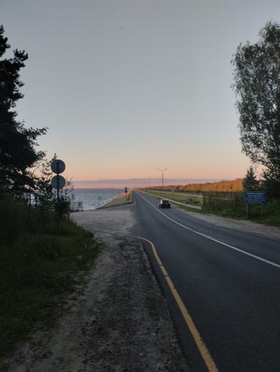 The image captures a scenic view of a highway running alongside a body of water, likely Lake Gorky, on the outskirts of Zavolzhye, Russia, during what appears to be sunset. The sky is a gradient of pale blue and soft orange, indicating the time of day.

On the left side of the frame, the dark silhouettes of trees dominate the foreground, with their branches reaching towards the sky. Adjacent to the trees is a dirt and gravel path, leading to the water's edge. Two circular traffic signs on a pole are visible near the water.

The highway, paved with asphalt, stretches into the distance, marked by white lines. A single black car is driving on the right lane. The left side of the highway is bordered by a fence and a line of trees, with a blue informational signpost visible in the distance on the right side of the road. The water is calm, reflecting the soft hues of the sky. The overall atmosphere is serene and quiet, typical of a rural highway at dusk.