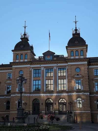 The image shows the Oulu City Hall, a grand, historical building with a light brown facade. The building features a central structure with two symmetrical towers topped with ornate spires. A flag pole with a black and white flag is visible between the towers. The facade is adorned with numerous windows, many of which have arched tops or intricate detailing. A lamppost stands in front of the building, and steps lead up to the main entrance. The scene is set against a clear blue sky, and the lower part of the image shows a landscaped area with flowers and foliage.