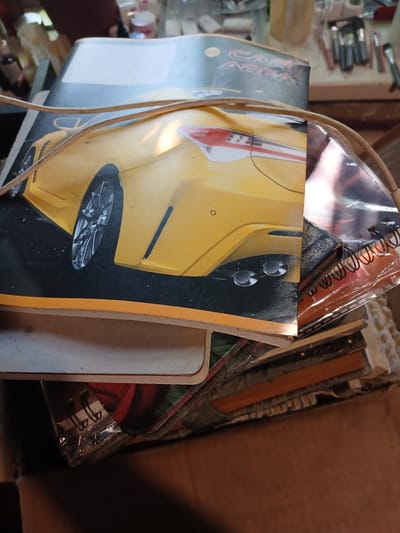 A notebook with a yellow sports car on the cover is partially visible, along with a tangle of wires and other notebooks. The items are stacked in a cardboard box. The scene is indoors, with a blurred background that suggests a cluttered environment with various objects on shelves, including what appear to be makeup brushes and containers. The lighting is dim and casts shadows. The location is identified as Chutove, Ukraine.