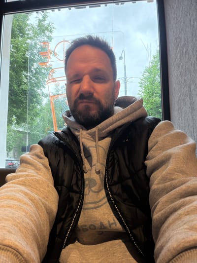 A man is seen indoors, near a window. Outside, there are trees and a street scene. Orange neon lettering can be seen through the glass. The man wears a gray hooded sweatshirt and a black vest. The text "URBAN MODERNURBAN" is visible on the vest. The location is Metallostroy, Russia.