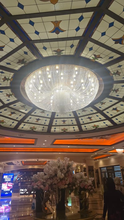 The image shows the interior of a building, likely a casino or hotel lobby, in Kazafani, Northern Cyprus. A large, ornate crystal chandelier hangs from a stained-glass ceiling. The ceiling features a geometric pattern with blue diamonds and floral motifs in yellow and green. Below the ceiling, there are brightly lit orange architectural elements. In the foreground, a large floral arrangement is displayed on a circular table with reflective surfaces. To the left, a digital screen advertises a "ROULETTE TOURNAMENT." In the background, there are entrances to various establishments, including one labeled "LUXURY BRAND." Several individuals are visible in the background, including two women facing away from the camera. The overall impression is one of opulence and high-end entertainment.