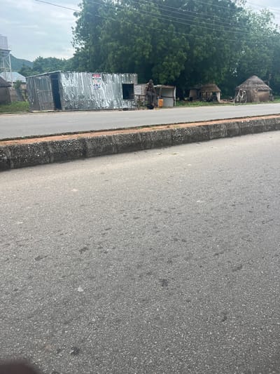 The image depicts a roadside scene in Kona Garu, Nigeria. The foreground is dominated by a paved road, with a concrete curb and a strip of earth and vegetation separating it from the background. In the background, there are several structures, including what appears to be a corrugated metal building, possibly a shop or dwelling, and some traditional thatched-roof huts. Lush green trees form a dense backdrop.
Visible elements include a bicycle leaning against one of the structures, and what might be a person sitting in front of the metal building. A yellow container or jerrycan is also visible near the metal structure. The overall scene suggests a rural or semi-rural setting, potentially a small village or roadside settlement.
The weather appears to be overcast, with a diffused light suggesting it might be morning or late afternoon, or simply a cloudy day. There is no explicit indication of a specific event or activity taking place, beyond the general presence of settlement and a road.
On the corrugated metal building, there is visible text, including a logo with the letters "POS" prominently displayed, along with other text in an illegible font. This suggests the building may be a point of sale or a service provider. The image is oriented vertically, with the road running diagonally from the bottom right to the top left.