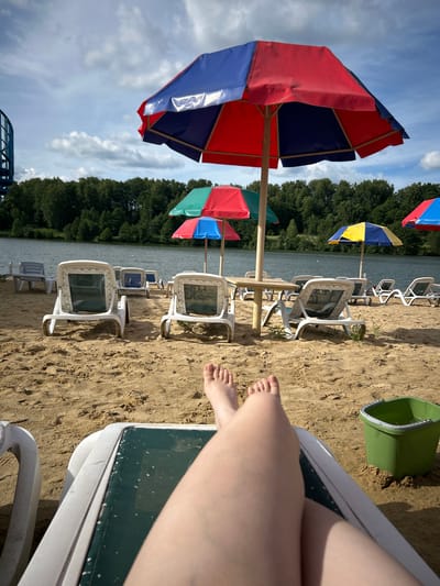 The image depicts a sun-drenched sandy beach adjacent to a calm body of water, likely a lake, bordered by a dense line of trees. Multiple rows of white plastic lounge chairs and colorful beach umbrellas are arranged on the sand, suggesting a public recreational area. In the foreground, a pair of bare feet are visible, resting on a green and white striped lounge chair. To the right of the chair, a bright green plastic bucket is partially filled with water and sand, placed on the beach. The sky is partly cloudy, with diffused sunlight casting shadows on the sand and illuminating the scene. The overall impression is one of a tranquil summer day at a beach resort, potentially located in or near the city of Yubileynyy, Russia, given the geographical context provided in the prompt.