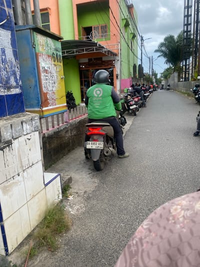 The image depicts a street scene in Sungai Paring, Indonesia. In the foreground, a person wearing a green Gojek uniform is seated on a scooter, facing away from the camera. The scooter has a license plate that reads "DA 6952 AEZ". The person is wearing a helmet. To the left of the scooter, there is a blue and yellow electrical box with faded markings and advertisements. Behind the electrical box, a building with orange and green facades is visible. Along the right side of the street, a row of parked scooters is lined up. The street is paved and appears to be a two-way road. In the background, several palm trees and a cloudy sky can be seen. The overall environment suggests a typical urban street in Indonesia, possibly a waiting area or a hub for ride-sharing services like Gojek.