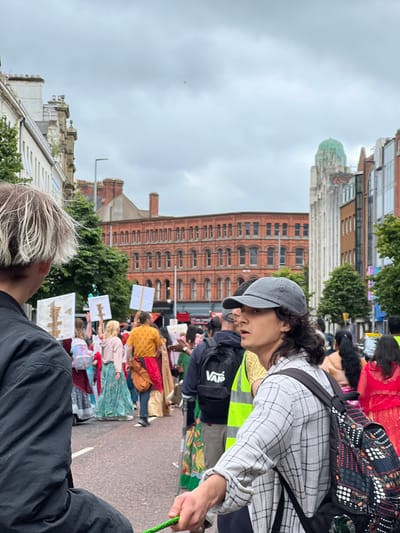 In Belfast City District, United Kingdom, a street scene unfolds under a cloudy sky. A diverse crowd is gathered, many holding white signs with symbols. In the foreground, two individuals are prominent: one with distinctive light-colored hair, seen from the back, wearing a black jacket; and another, facing towards the right, wearing a grey baseball cap and a plaid shirt, with a backpack adorned with a patterned design. Behind them, a larger group of people are visible, some in colorful attire, walking along a street lined with buildings. The architecture suggests an urban environment, with a prominent red brick building with multiple windows and a distinctive green dome structure in the distance. The ground is paved, indicating a public thoroughfare. The overall impression is of a gathering or procession in a city setting.