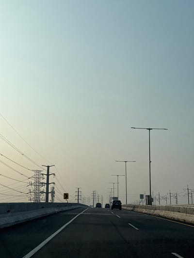 The image shows an elevated highway in Gandamekar, Indonesia. The road surface is dark asphalt with white lane markings. Several vehicles are visible in the distance traveling in the same direction. Streetlights line the edge of the road. Beyond the concrete barrier, power lines and electrical towers are present. The sky is a pale blue-gray. A yellow traffic sign is visible next to the road. The image is rotated 90 degrees counterclockwise.
