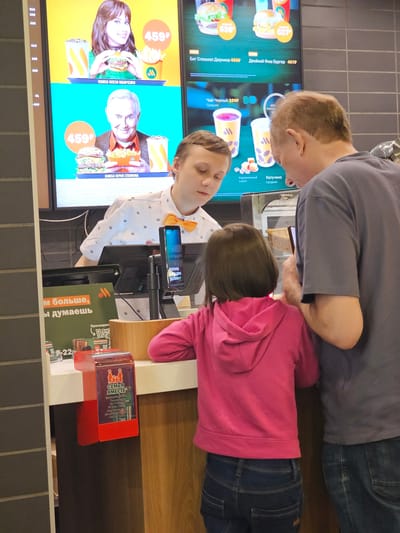 The scene depicts the interior of a fast-food restaurant, likely in Mozhaisk, Russia. A man and a child, both facing the counter, are making an order. The man is wearing a gray shirt and jeans, while the child has on a pink hoodie and jeans. A worker wearing a white shirt with a yellow bow tie is behind the counter, possibly taking their order. On the left, there is a digital menu board displaying food items and prices, including "Биг Спешиал Джуниор," "Двойной Фиш Бургер," "Чай Черный," and "Капучино." Promotional images feature personalities Олеся Иванченко and Юрий Стоянов. The counter area has a point-of-sale system with a screen showing "платите как вам удобно". The text "м больше, ы думаешь" is printed on a sign. A red promotional display stands on the counter. The background has a wall tiled with gray, rectangular tiles.