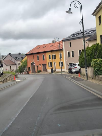 The image captures a street scene in Weiler-la-Tour, Luxembourg, on an overcast day. The road curves to the left, with a slightly wet asphalt surface marked by faint white lines. On the right side of the road, a light-colored concrete wall is topped with dense green hedges and a chain-link fence. Two cars are parked on the right: a black SUV behind a white SUV. A traffic cone is visible on the sidewalk near the white SUV.
The left side of the street features a stone retaining wall and a partially visible white vehicle. Orange traffic cones and construction barriers suggest ongoing road work or maintenance. Further down the street, a row of multi-story buildings with terracotta roofs lines the right side. These buildings have distinct architectural styles, with varying colors of facades, including orange, yellow, and beige. Several streetlights with decorative, spiral-shaped posts are positioned along the sidewalk. The sky is filled with heavy, gray clouds.