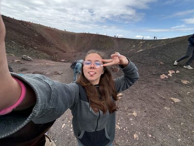 A person wearing glasses, a gray hooded jacket, and jeans is taking a selfie on Mount Etna in Nicolosi, Italy. They are holding up a peace sign and sticking their tongue out. The background consists of a dark, rocky terrain with other people visible in the distance and a sky filled with clouds.