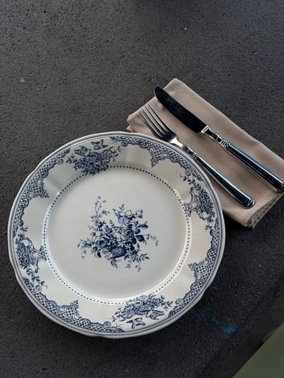 A place setting for one, consisting of a white plate with a blue floral pattern and a silver fork and knife resting on a beige napkin, is arranged on a dark, textured surface. The plate is centrally positioned, with the cutlery situated to its upper right. The background is dark and appears to be an outdoor or industrial surface, with subtle blue smudges visible in the lower right quadrant. The lighting suggests an overhead source, creating soft shadows. No people are present in the image. The scene is likely an outdoor dining setup in Dsegh, Armenia, potentially in a restaurant or a picturesque outdoor location, given the naturalistic setting suggested by the plate's design and the dark, stone-like surface.