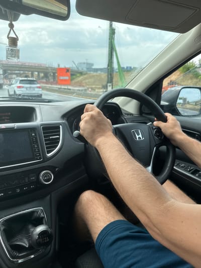 A person is driving a Honda car in Diegem, Belgium. The driver is gripping the steering wheel with both hands. The car's dashboard, infotainment system, air vents, climate control buttons, and gear stick are visible. Outside the car window, a construction site is apparent, featuring an orange sign and building construction cranes. Other cars are driving on the road. The weather appears overcast. An air freshener hangs from the rearview mirror.