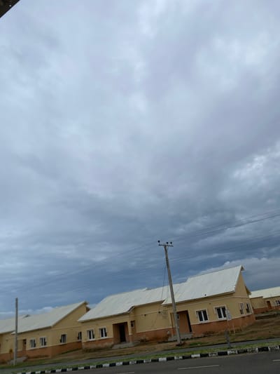The image shows a row of tan-colored houses with white roofs, likely in a residential development. The houses have multiple windows and appear to be two stories tall. A paved road with black and white markings runs alongside the houses, with a strip of dry grass and dirt in between. Overhead, power lines stretch across the sky, supported by utility poles. The sky is cloudy and appears overcast, suggesting a possibly dim or approaching rainy day. The overall scene depicts a suburban or semi-rural residential area. The orientation of the image is slightly tilted, with the buildings appearing to extend upwards from the bottom right towards the top left. There are no people visible in the image. The time of day is difficult to determine definitively due to the overcast conditions, but it appears to be daytime. There are no clear location cues besides the provided context of Dumnga, Nigeria. There is no visible text in the image.
