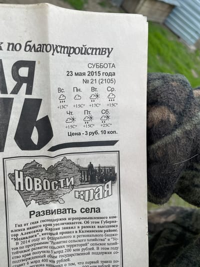 A close-up shot shows a section of a newspaper, presumably from Talmenka, Russia, dated Saturday, May 23, 2015. The headline of an article reads "Develop the Villages," accompanied by a map illustration of the region. Text details the increase in state support for the agro-industrial complex, mentioning Governor Alexander Karlin and the "Medialogue" event in the Kalmansky district. Temperature forecasts for the week are shown, ranging from +13°C to +25°C. The price is listed as 3 rubles and 10 kopecks. In the upper portion of the image, there is a blurred object that appears to be a gloved hand. The setting appears to be outdoors.