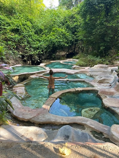 A woman with her back to the camera stands in a series of tiered, natural hot spring pools in Khlong Thom Nuea, Thailand. She is wearing a leopard-print bikini and has her arms outstretched. The pools are filled with clear turquoise water and are surrounded by natural rock formations and lush green vegetation. The scene is set amidst a dense tropical forest, with sunlight filtering through the trees.