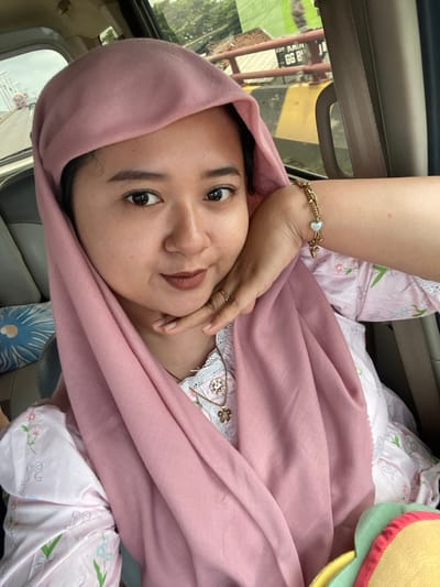 A woman with fair skin is wearing a pink headscarf and resting her hand on her face. She has dark eyes and eyebrows, and her lips are slightly parted. She is wearing a gold bracelet with a heart charm. The background suggests she is in a car, and she is wearing a pink outfit with a floral pattern. There is a plush, colorful object near her right shoulder.